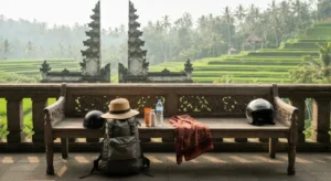 Plecak, kapelusz, kask i sarong na drewnianej lawie tarasu na Bali, w tle kamienna brama swiatyni i tarasy ryzowe