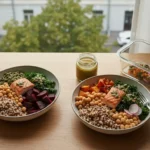 Trzy glebokie misy z warstwowym lunchem na drewnianym stole przy oknie kamienicy, obok szklany pojemnik na meal prep